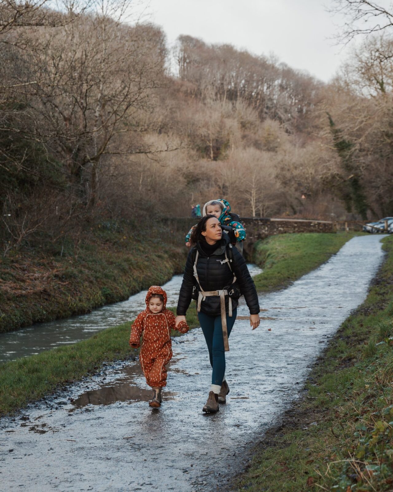 a mum walking with her baby on her back and holding the hand of her older little girl next to a river in cornwall